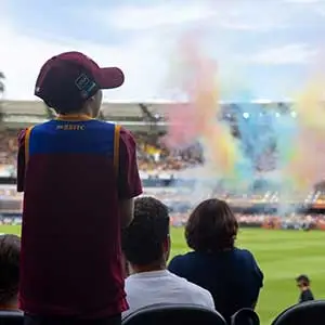 A young child in care, wearing a maroon and blue jersey, stands with hope and excitement in a stadium as colourful smoke fills the air—surrounded by supportive adults, symbolising inclusion, belonging, and brighter futures through the Empower Youth Program.